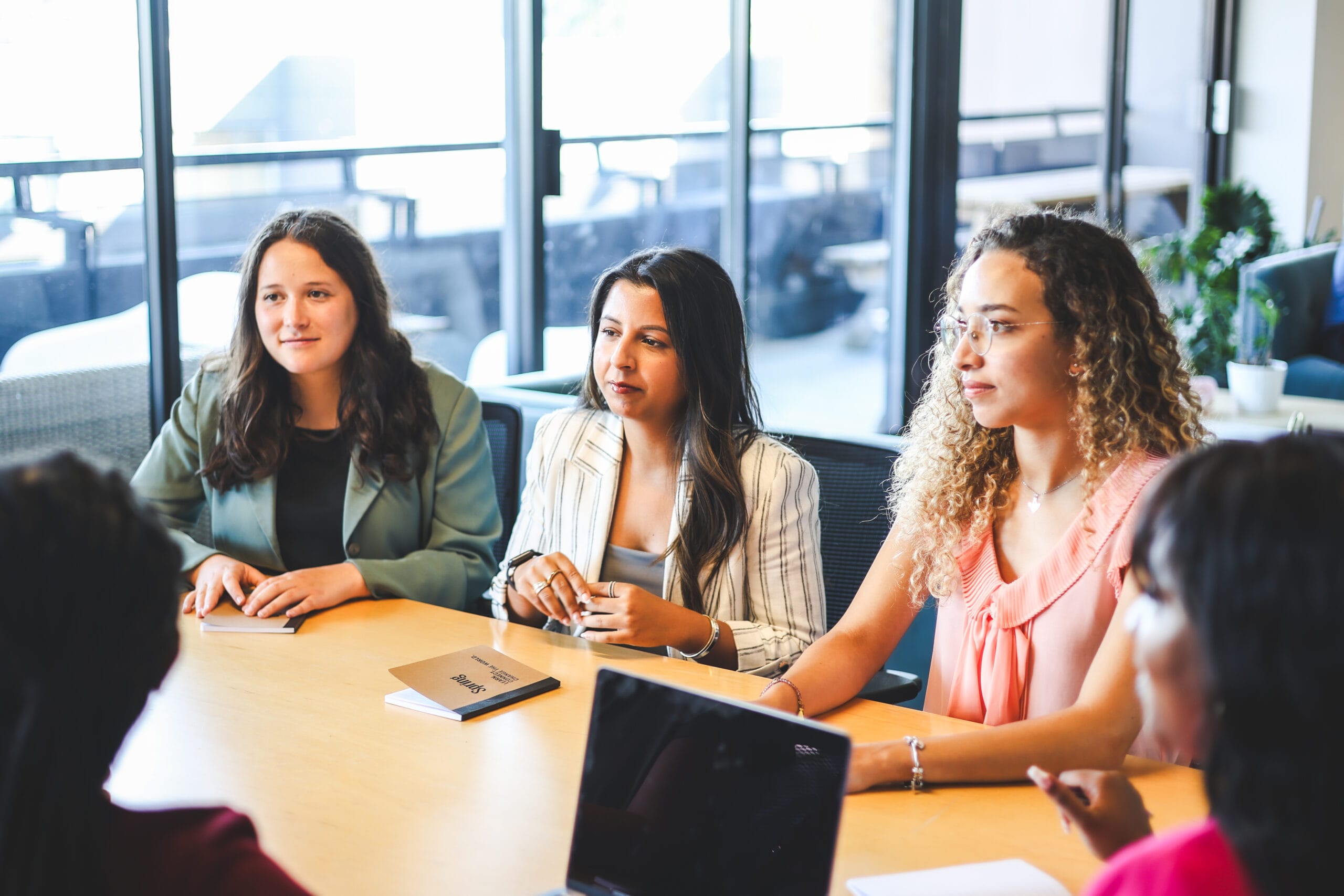 Three women participating in a meeting. A notebook with the title 'Spring' sits in the center of the table