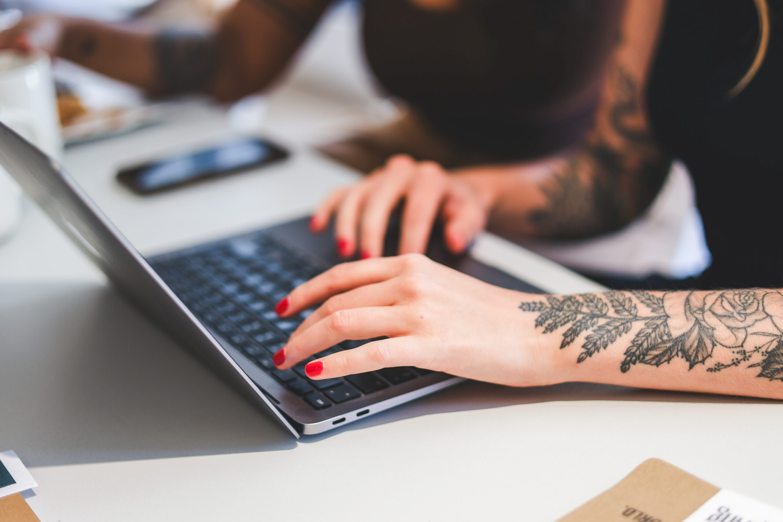 A cropped photograph of a woman working on a laptop computer. She has tattoos on both of her arms