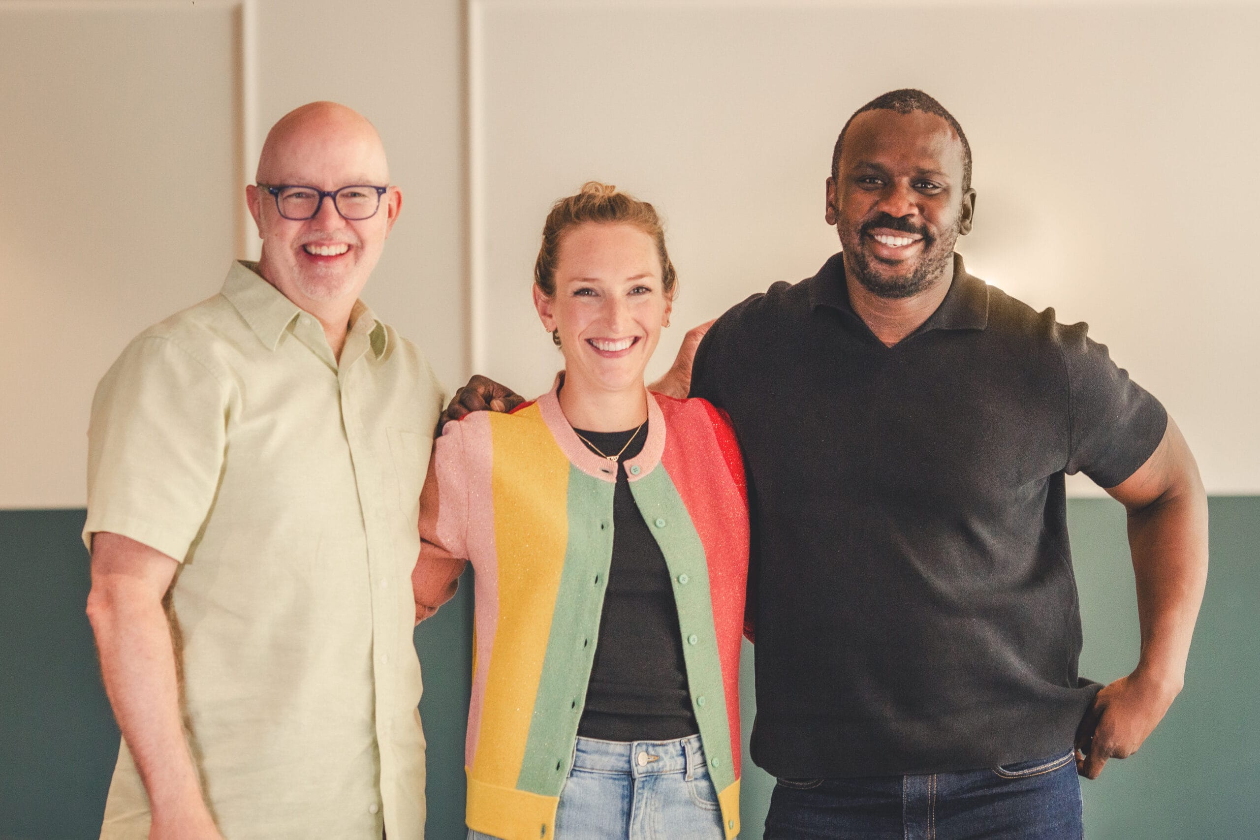 A man in a button up polo wearing glasses, a women in a striped cardigan and a man with a beard wearing a polo shirt with their arms behind eachother  all smile
