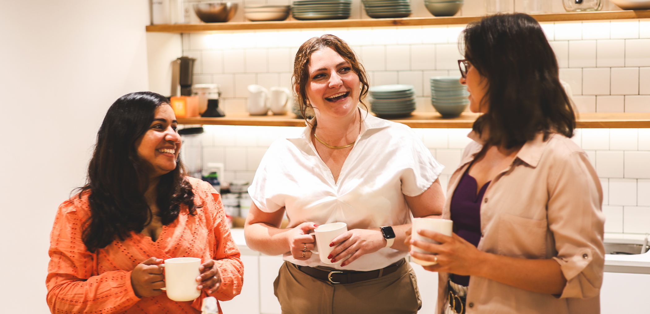 3 women stand together with coffee mugs in hand having a conversations