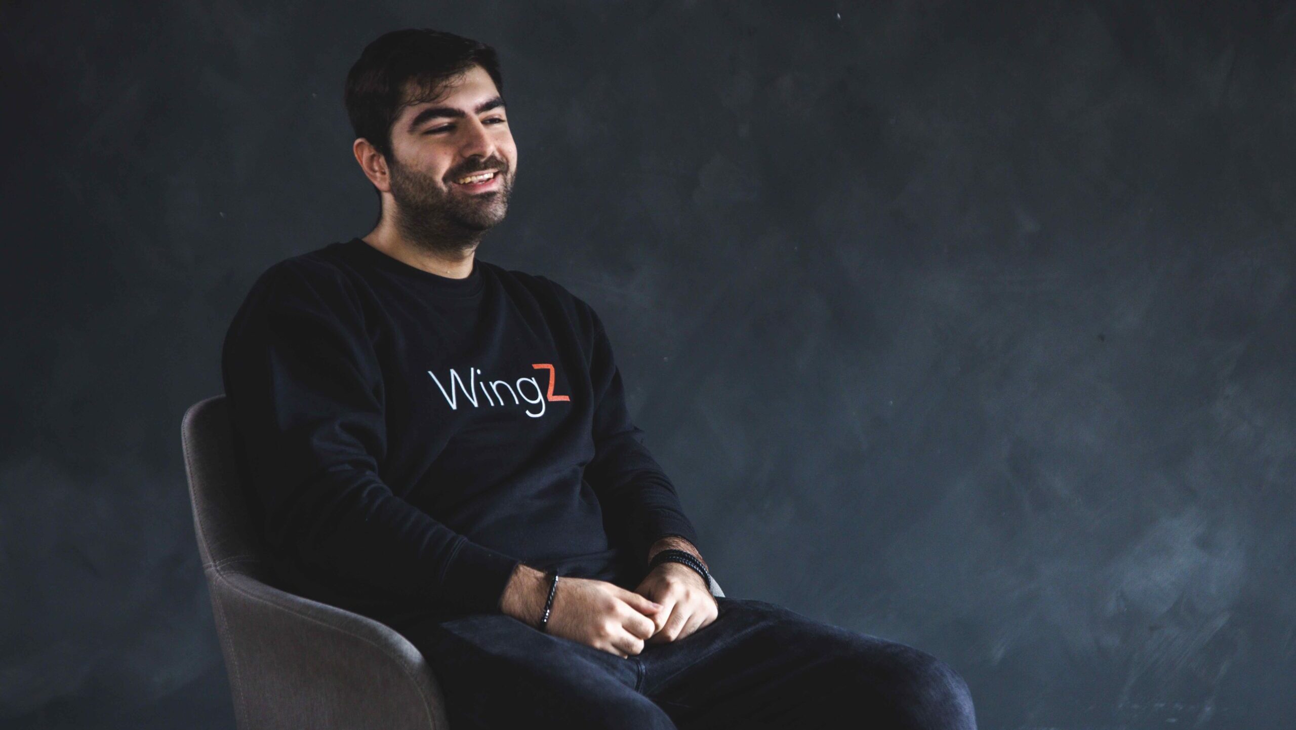 WingZ's Hossein Rezai, wearing a WingZ t-shirt, sitting in a chair against a dark grey backdrop and smiling