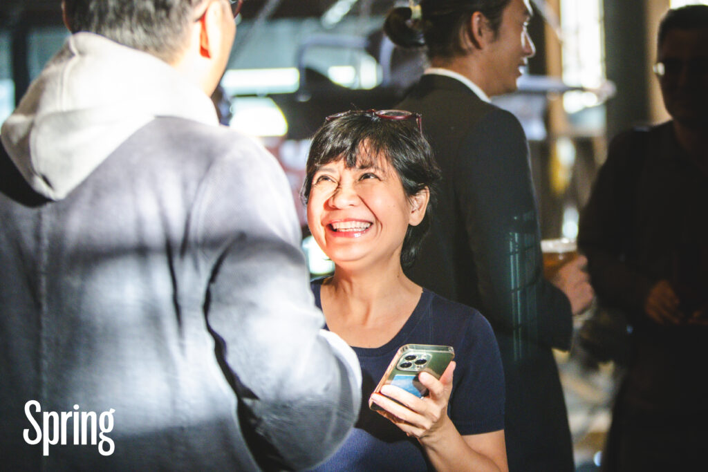 A person at a Spring event holding their phone, talking and smiling at a tall person with their back to the camera