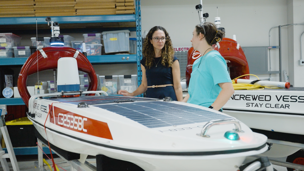 Two people, including CEO and Co-Founder Julie Angus of Open Ocean Robotics, stand in a workshop with several marine vessels.