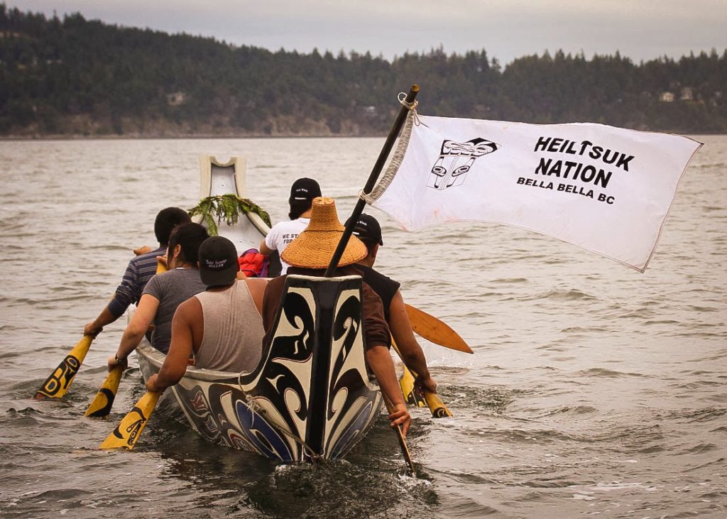 Rear shot of people paddling a painted canoe. The passenger in the rear is wearing a woven cedar hat, and the canoe bears a flag saying “Heiltsuk Nation Bella Bella BC.”