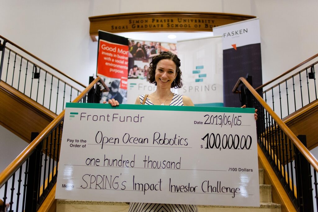 A smiling person holds a giant cheque awarding Open Ocean Robotics $100,000 as part of Spring’s Impact Investor Challenge.